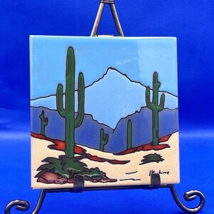 May include: Square ceramic tile depicting a desert scene with green cacti, a light blue sky, and mountains. The tile has a cream base with red and green details. The tile is displayed on a small metal easel.