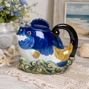 May include: A ceramic teapot shaped like a fish with a glossy deep blue upper body and a white underbelly. The handle is dark brown, and the base features sculpted green waves. The fish has a prominent dorsal fin lid and a gold-toned tail fin.