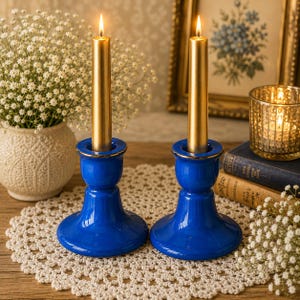 Puede incluir: Dos candelabros de cerámica azul cobalto brillante con bordes dorados, cada uno con una vela cónica de color oro metálico con una llama encendida. Los candelabros cuentan con una base ancha y estable y un diseño de cuello redondeado.