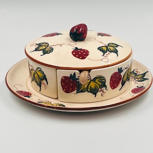 Op de afbeelding: Een crème-kleurige keramische boterpot met een deksel en bijpassende schotel. De pot en het deksel zijn versierd met rode aardbeien en groene bladeren. Het deksel heeft een aardbeivormige knop. De schotel heeft een rode rand.