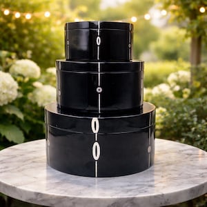 May include: A set of three black lacquered storage boxes stacked on a marble table. Each box has a white vertical line with the number "0" and small circles. The boxes are different sizes and have a glossy finish.