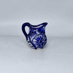 May include: A small, blue ceramic pitcher with a handle and a spout. The pitcher is decorated with a floral pattern in white and blue. The pitcher is approximately 5 inches tall.