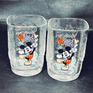 Puede incluir: Dos vasos cuadrados transparentes con un diseño texturizado, cada uno con Mickey Mouse. Los vasos tienen un diseño colorido con fuegos artificiales y el año "2000". Ideales para servir bebidas.