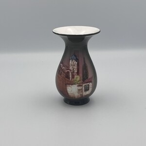 May include: A decorative vase with a dark brown body and a white rim. The vase features a painted scene of a building with a blue roof and a white facade. The vase is a decorative item.