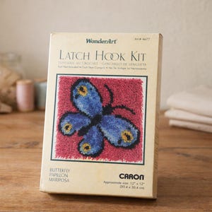 May include: A WonderArt latch hook kit featuring a butterfly design. The kit includes a canvas with a red background and a blue butterfly with yellow accents. The approximate size is 12" x 12".