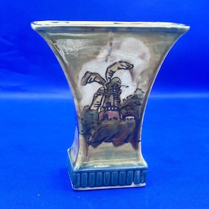 May include: A decorative vase with a flared top and a square base. The vase features a landscape scene with a windmill and buildings in shades of brown, green, and blue. The base has a blue, ribbed design.