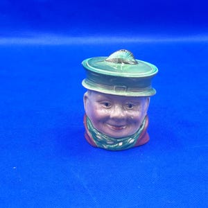 Könnte beinhalten: Keramikfigur eines lächelnden Männerkopfes mit grünem Hut und Schal. Das Gesicht ist mit rosigen Wangen und einem roten Kragen bemalt. Der Hut hat ein Muscheldesign auf der Oberseite. Der Hintergrund ist einfarbig blau.