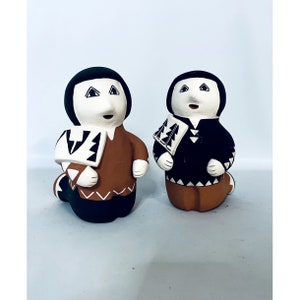 May include: Two ceramic figurines of people kneeling, wearing traditional clothing with geometric patterns in black, white, and brown. The figurines are made of terracotta clay and have a rustic, handmade look.
