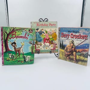 Puede incluir: Tres libros infantiles antiguos. Las portadas presentan ilustraciones coloridas de Bambi, una escena de fiesta de cumpleaños y Davy Crockett. Los libros se exhiben sobre una superficie blanca.