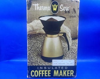 Puede incluir: Una cafetera aislada en su caja original. La caja presenta la marca "Thermo Serv" y una cafetera dorada y negra. La cafetera tiene un portafiltro negro y una jarra dorada. La caja está sobre un fondo azul.
