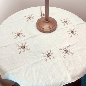 May include: A white, round tablecloth with embroidered snowflake-like designs in brown and tan. A brown lamp base and stem are centered on the cloth. The tablecloth is on a round table.