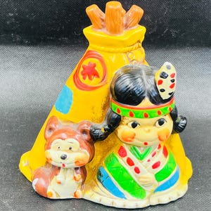 Può includere: Una statuetta in ceramica raffigurante una ragazza nativa americana, un piccolo animale e un teepee giallo. La ragazza indossa un abito colorato e un copricapo di piume. Il teepee ha un design a stella rossa e accenti blu. La statuetta è dipinta con colori vivaci.