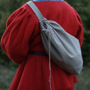 May include: A gray canvas shoulder bag with a drawstring closure, worn over a red coat. The bag has a long strap and a braided cord. The coat has large sleeves and a black belt. The background is blurred green.