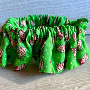 May include: A bright green fabric scrunchie featuring a repeating pattern of white and red striped candy canes tied with green bows, accented by small white dots scattered across the material, creating a gathered, ruffled circular shape.