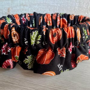 May include: A circular fabric bowl cover featuring a black background with a repeating pattern of orange pumpkins and green autumn leaves. The cover has an elasticized edge that creates a gathered, ruffled appearance around the perimeter.