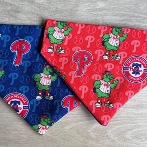 May include: Two dog bandanas, one blue and one red, with a Phillies baseball team logo and mascot. The bandanas are made of fabric and have a triangular shape.