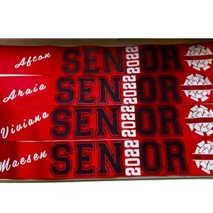 以下が含まれることがあります： 白文字で「Senior 2022」と書かれた、赤と黒の布製のサッシュが4本。サッシュには、「Afton」、「Araia」、「Viviana」、「Maesen」の名前が印刷されています。