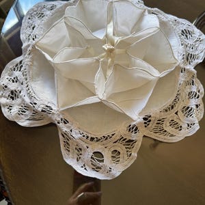 Puede incluir: Un recipiente blanco, de estilo vintage, con forma de flor y múltiples compartimentos. El recipiente está hecho de tela blanca y presenta un delicado ribete de encaje en los bordes. Una cinta de raso está atada en el centro.