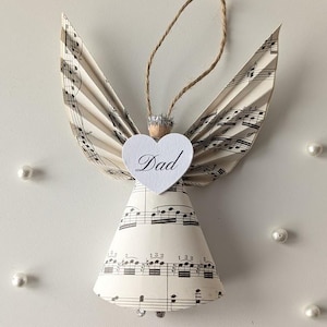 以下が含まれることがあります： 楽譜で作られた手作りの天使のオーナメント。天使は紙の羽、円錐形の体、そして「Dad」という言葉が書かれたハート型の顔をしています。吊り下げ用の紐ループが付いています。オーナメントは銀色のグリッターで飾られています。