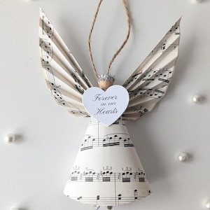 以下が含まれることがあります： 楽譜で作られた手作りの天使のオーナメント。天使は紙の羽と円錐形の体を持っています。「Forever in our Hearts」という言葉が書かれたハート型のタグが付いています。吊り下げ用の紐ループが上部にあります。