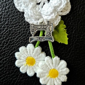 May include: White crocheted flowers with a silver sewing machine charm and green ribbon accents.