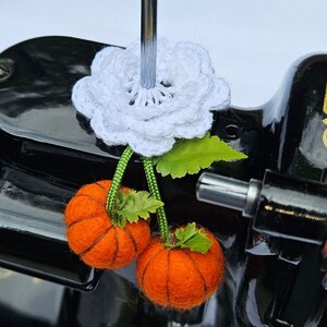 May include: A white crocheted flower with green leaves and two orange felt pumpkins hanging from it. The flower and pumpkins are attached to a black sewing machine.