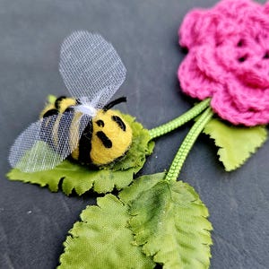 May include: A felt bee with black and yellow stripes and a mesh wing sits on a green leaf. The bee is next to a pink crocheted flower with green leaves and stems.