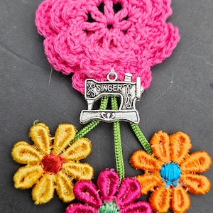 May include: A handmade floral brooch featuring a large, bright pink crocheted flower. Below, three embroidered flowers in yellow, orange, and pink are attached. A silver charm with the word "SINGER" and a sewing machine design hangs from the large flower.