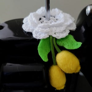 May include: A white crocheted flower with green felt leaves and yellow felt lemons attached to the stem. The flower and lemons are attached to a black sewing machine.