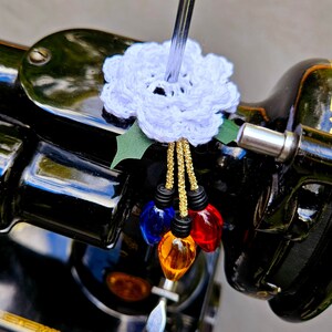 May include: A handmade Christmas ornament featuring a white crocheted flower with green leaves and gold string. Below the flower are four colorful light bulb ornaments in red, blue, and amber colors. The ornament is attached to a black object.