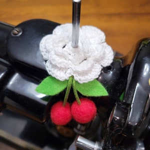 May include: A white crocheted flower with green felt leaves and two red felt cherries attached to the bottom. The flower is attached to a metal rod.