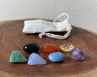 May include: Seven chakra stones in a variety of colors, including black, green, blue, orange, yellow, and purple. The stones are arranged in a circle on a wooden surface.