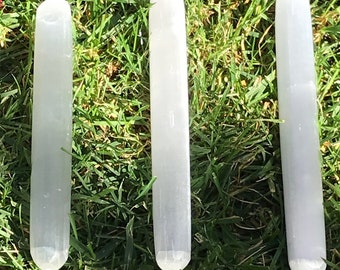 May include: Three white selenite wands, each with a slightly different shape and size, are arranged in a row on a bed of green grass. The wands are smooth and polished, with a slightly translucent appearance. The numbers 2, 3, and 4 are written below each wand.