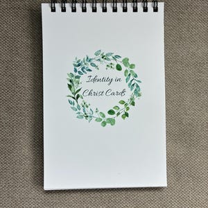 May include: A white spiral-bound notepad with the text "Identity in Christ Cards" inside a green wreath design. The notepad is on a neutral-colored surface.