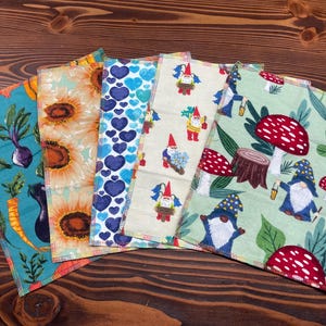 May include: Five rectangular fabric cloths featuring distinct patterns: a teal cloth with vegetables, a sunflower print, a blue heart pattern, a cream cloth with gnomes, and a light green cloth with gnomes, mushrooms, and tree stumps.