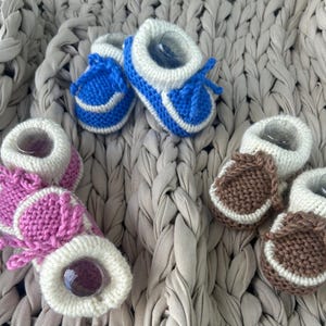 May include: A collection of handmade baby booties in blue, pink, and brown. The booties are knitted with white trim and a soft, plush interior. The blue booties have white accents, the pink booties have white accents, and the brown booties have white accents.