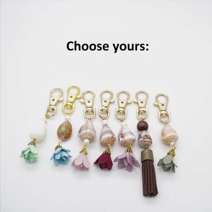 May include: Eight keychains with different charms. Each keychain has a gold metal clasp and a variety of charms, including seashells, tassels, and fabric flowers in shades of blue, green, purple, and brown.