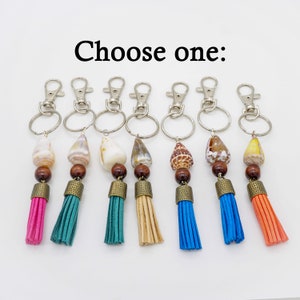 May include: Choose one: Seven keychains with different colored tassels and seashell charms. The tassels are pink, teal, teal, beige, blue, blue, and orange. The seashells are white, brown, brown, brown, brown, brown, and yellow.