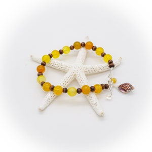May include: A beaded bracelet featuring yellow and orange gemstone beads interspersed with dark wooden beads. A silver charm with a seashell and a clear crystal hangs from the bracelet. The bracelet is displayed on a white starfish.