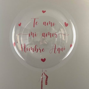 Op de afbeelding: Een doorzichtige bubbelballon met rode tekst "Te amo mi amor Nombre Aqui" en kleine rode hartaccenten. De ballon is vastgebonden met een rood lint.