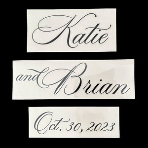 May include: Three white cards with black calligraphy text. The top card says "Katie". The middle card says "and Brian". The bottom card says "Oct. 30, 2023".