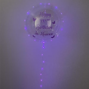 May include: A clear, round balloon with purple LED lights and the text "Happy XXnd Birthday In Heaven." The balloon features butterfly graphics and a string of lights extending downward.