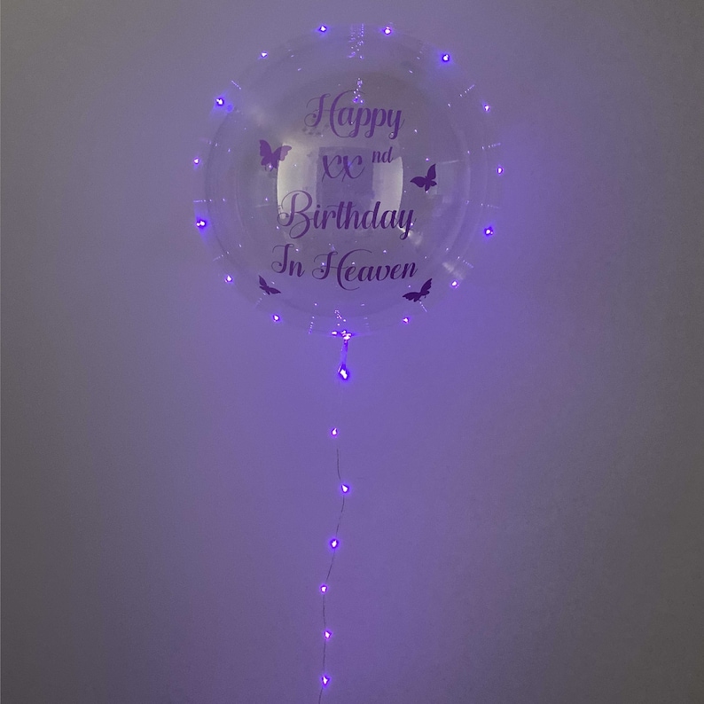 May include: A clear, round balloon with purple LED lights and the text "Happy XXnd Birthday In Heaven." The balloon features butterfly graphics and a string of lights extending downward.