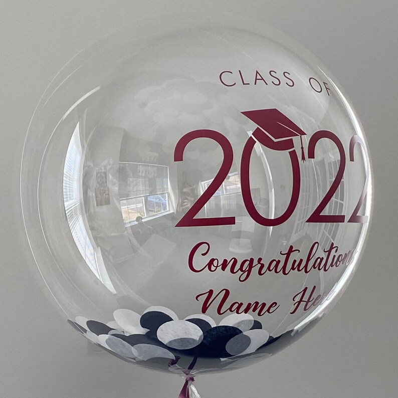 May include: A transparent bubble balloon with maroon text reading "CLASS OF 2022 Congratulations Name Here". The balloon features a graduation cap graphic and is filled with white and navy blue confetti.