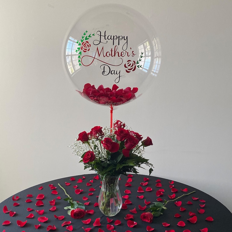 Peut inclure: Un ballon transparent rempli de p&eacute;tales de roses rouges avec le texte "Happy Mother's Day" imprim&eacute; dessus. Un bouquet de roses rouges dans un vase en verre transparent se trouve sous le ballon. Des p&eacute;tales de roses rouges sont &eacute;parpill&eacute;s sur une nappe de table noire.