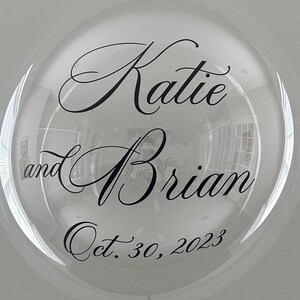 May include: Clear balloon with black calligraphy lettering that reads "Katie and Brian Oct. 30, 2023".