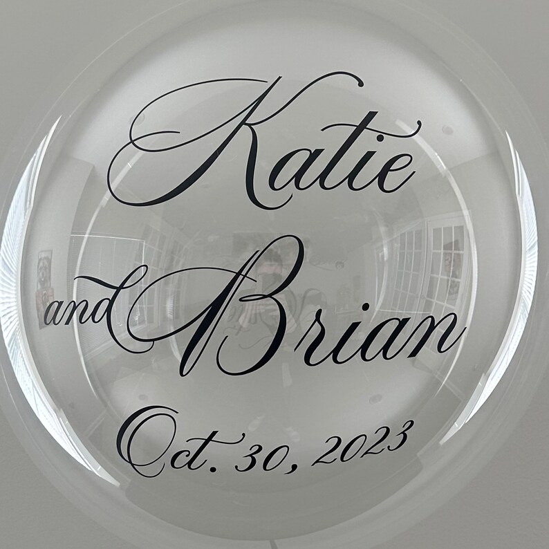 May include: Clear balloon with black calligraphy lettering that reads "Katie and Brian Oct. 30, 2023".