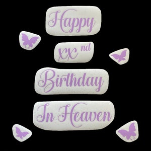 May include: A collection of white, textured stones with purple script that reads "Happy", "XX nd", "Birthday", and "In Heaven". Several small butterfly-shaped stones in purple are scattered around the main stones. The background is black.