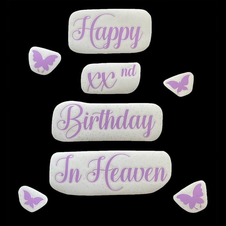 May include: A collection of white, textured stones with purple script that reads "Happy", "XX nd", "Birthday", and "In Heaven". Several small butterfly-shaped stones in purple are scattered around the main stones. The background is black.