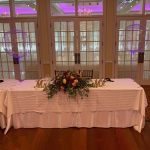 May include: A white tablecloth with a striped pattern covers a table with a large floral centerpiece. The table is set for two with the words "Mr." and "Mrs." in wooden letters. Two clear balloons with purple lights are in the background.
